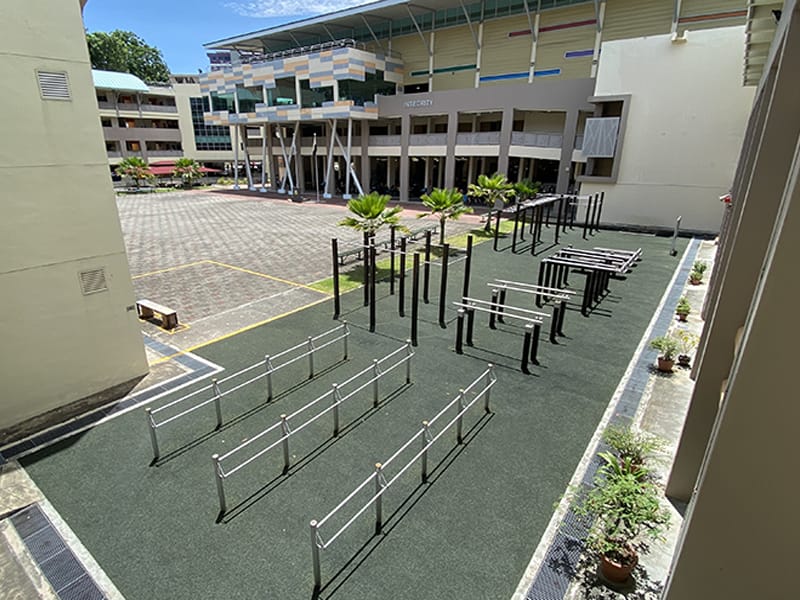 Outdoor fitness area with metal bars and parallel bars on green turf beside a multi-story school building.